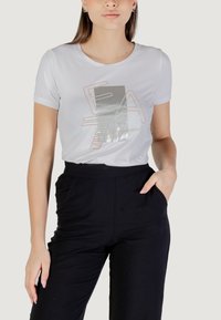 Camiseta de algodón blanca de manga corta, con un diseño gráfico plateado y detalles en cobre. Combinada con pantalones negros de talle alto.