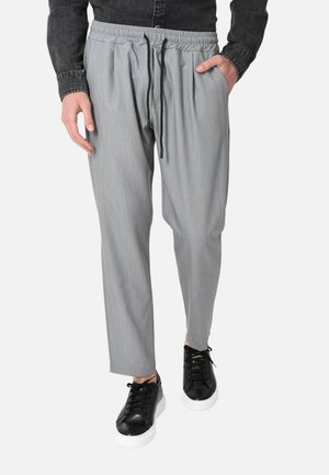 Persona che indossa pantaloni grigi chiaro con coulisse e scarpe da ginnastica nere con suole bianche, mani nelle tasche, in piedi davanti a uno sfondo neutro.