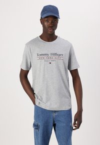 Grå bomulds t-shirt med "Tommy Hilfiger" logo i marineblå og rød. Kombineret med lyseblå cargo jeans og en marineblå kasket.