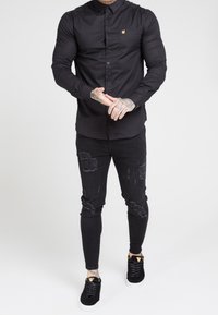 Musta kauluksellinen nappipaita, yhdistetty kuluneisiin mustiin kapealahkeisiin farkkuihin ja mustiin lenkkareihin, joissa on kultaiset yksityiskohdat.