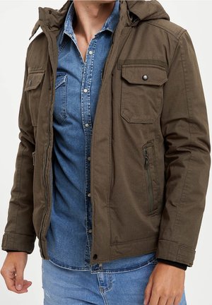 Uomo che indossa una giacca verde oliva sopra una camicia di jeans blu con bottoni e jeans azzurri, mostrato dal collo ai fianchi.