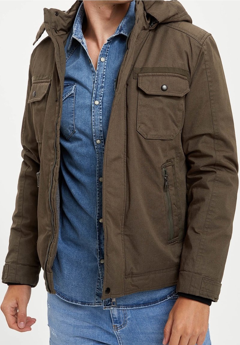 Mężczyzna ubrany w oliwkowozieloną kurtkę na niebieską dżinsową koszulę zapinaną na guziki oraz jasnoniebieskie jeansy, widoczny od szyi do bioder.