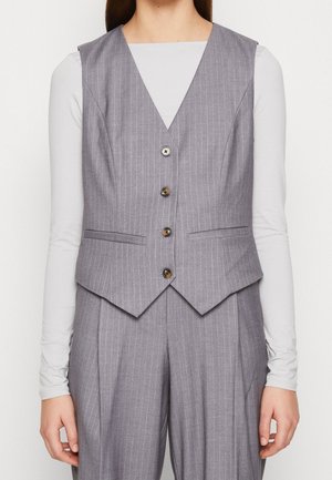 Femme portant un gilet gris à fines rayures et un pantalon taille haute assorti sur un haut blanc à manches longues, debout, les bras détendus.