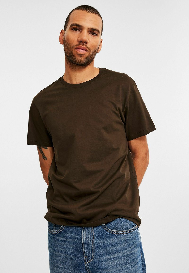 Mand med kort hår og skæg iført en mørkebrun T-shirt og blå jeans, stående med hænderne bag ryggen mod en hvid baggrund.