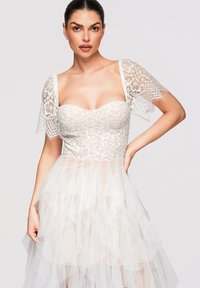 Femme portant une robe blanche bustier en dentelle avec des manches courtes en dentelle florale et une jupe en tulle à plusieurs couches, posant avec une main sur la hanche.