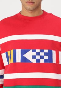 Sarkana sporta krekla ar horizontālām baltām svītrām un zilu, dzeltenu un baltu rakstu joslu krūšu daļā. Ribotā apkakle un apakšmala.