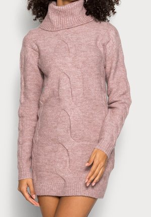 Femme portant une robe pull-col roulé en maille torsadée rose pâle avec des manches longues, debout les bras détendus le long du corps.