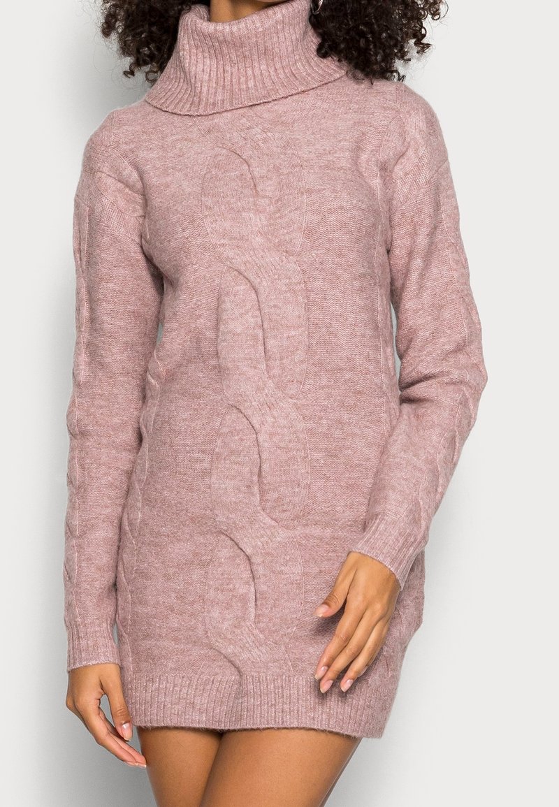 Femme portant une robe pull-col roulé en maille torsadée rose pâle avec des manches longues, debout les bras détendus le long du corps.