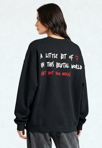 Fekete, oversized kapucnis pulóver, fehér és piros felirattal a háton, kerek nyakú és bordás mandzsettákkal, puha anyagból készült.