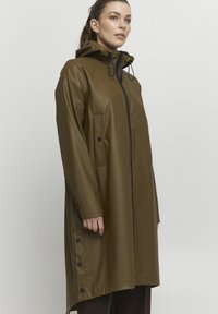 Trenchcoat lung de culoare verde măsliniu, realizat din material impermeabil, cu glugă. Dispune de închidere cu fermoar în față, două buzunare laterale și detalii cu capse la tiv.