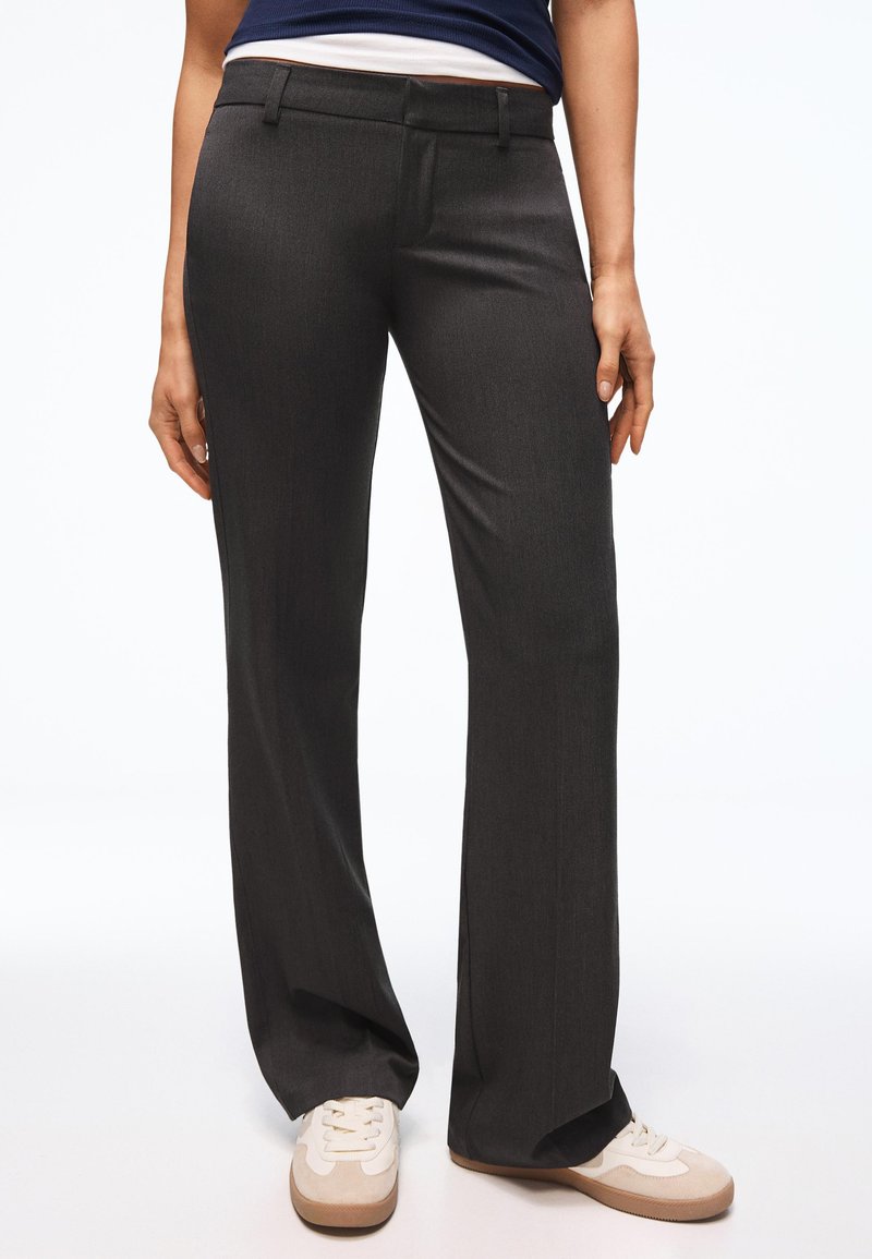 Femme portant un pantalon droit gris foncé associé à des baskets beige et blanches, debout devant un fond blanc uni.