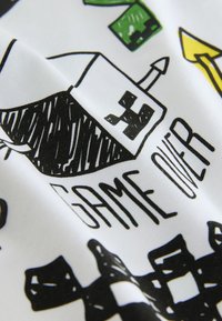 Un personnage pixellisé noir et blanc avec une tête carrée et une antenne au-dessus des mots "GAME OVER" sur un tissu blanc.
