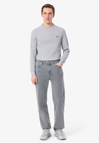 Chemise henley à manches longues gris avec patte de boutonnage à trois boutons, portée avec un jean taille haute gris clair et des baskets grises, présentant un logo subtil.