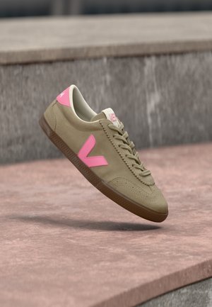 Beige sneaker met roze logo en hielstuk, bruine zool, zwevend boven een roze stenen oppervlak en een grijze betonachtergrond.