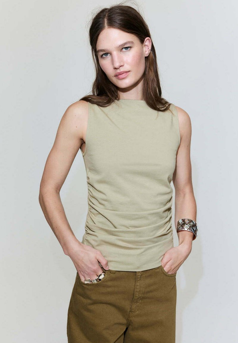 Jeune femme portant un haut beige sans manches et un pantalon marron, les mains dans les poches, sur un fond clair uni.