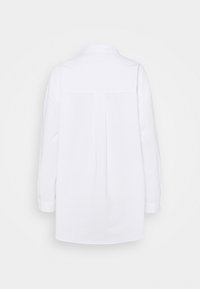 Chemise blanche à manches longues avec un pli au dos, poignets boutonnés et col classique, vue de dos sur un fond uni.