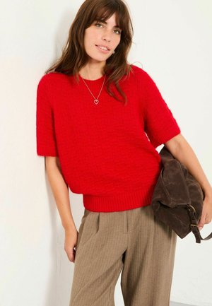 Red textured knit sweater with short sleeves and ribbed hem, paired with beige pleated trousers. Dark brown shoulder bag.