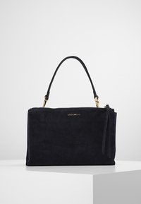 Czarna zamszowa torebka na krótkim uchwycie, ze złotymi elementami i logo COCCINELLE z przodu, umieszczona na białym podestie na jasnym tle.