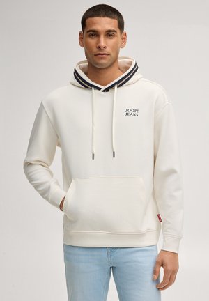 Mężczyzna w kremowej bluzie z kapturem z napisem "JOOP! JEANS", jasnoniebieskich dżinsach, stojący z jedną ręką w kieszeni na jasnym tle.