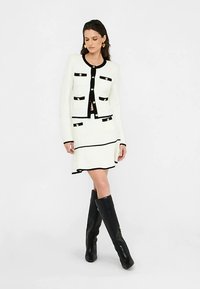 Balta megzta kardiganas su juodais akcentais ir auksiniais mygtukais, derinamas su balta suknele su juodos linijos dizainu. Modelis vilki juodus, iki kelių siekiančius batus.