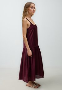 Abito maxi senza maniche color bordeaux con corpetto aderente e gonna a campana arricciata. Il tessuto sembra leggero con una texture liscia.