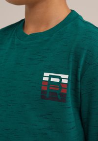 Grön kortärmad T-shirt med en texturerad yta. Har en frampatch med logotyp i vitt, rött och svarta horisontella ränder. Rund halsringning.