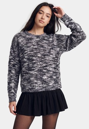Jonge vrouw met lang donker haar, die een zwart-grijze gebreide trui en een korte zwarte rok draagt tegen een witte achtergrond.