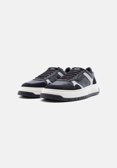 Paire de baskets noires et grises perforées avec des semelles blanches épaisses et des lacets noirs, présentées en angle pour montrer les vues de côté et de face.