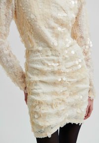 Zbliżenie sukienki pokrytej cekinami w kolorze kremowym, z długimi rękawami i marszczoną talią, noszonej z czarnymi rajstopami na jasnym tle.
