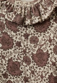 Virágmintás szövet, amely bonyolult bordó virágokat és indákat tartalmaz egy krém színű háttéren, fodros nyakkivágással.