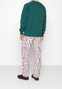 Sudadera verde con cuello redondo clásico, combinada con pantalones rosas que presentan un divertido patrón de caras. Un calzado negro completa el atuendo.