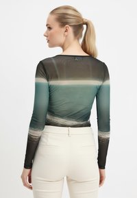 Maglietta a maniche lunghe in tessuto trasparente con design a gradiente, caratterizzata da tonalità di verde e colori terra, abbinata a pantaloni color crema.