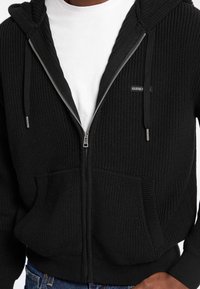 Sudadera con cremallera negra de punto con una superficie texturizada, bolsillos frontales y capucha con cordón. Presenta un pequeño logotipo en el pecho.