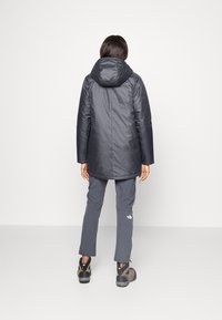 Czarna izolowana kurtka z kapturem, o gładkiej fakturze i dłuższym kroju. Noszona z szarymi spodniami i butami trekkingowymi, widok z tyłu.