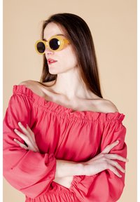 Blusa corallo off-the-shoulder con scollatura arricciata e maniche a palloncino, abbinata a occhiali da sole gialli rotondi con lenti scure.