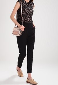 Top sin mangas de encaje negro, pantalones negros de cintura alta y zapatos de cordones en un beige claro. Un bolso cruzado metálico añade un acento reflectante.
