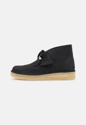 Bottine en daim noir avec semelle en crêpe, fermée par des lacets à l'avant, détails cousus, étiquette sur le côté avec la marque "Clarks Originals".