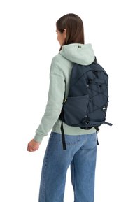 Marineblauwe stoffen rugzak met verstelbare schouderbanden, een voorvak en bungee koorden. Draagbaar over een lichtgroene hoodie en blauwe jeans.