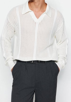 Man draagt een wit gebreide polotrui met lange mouwen, ingestopt in donkergrijze geplooide broek, handen in de zakken.