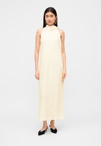 Μπλουζοφόρεμα σε κίτρινο χρώμα χωρίς μανίκια, με ψηλό λαιμό και λεπτομέρεια με πτυχώσεις, από λείο ύφασμα και με μήκος μέχρι το πάτωμα. Συνδυασμένο με μαύρα μυτερά παπούτσια.