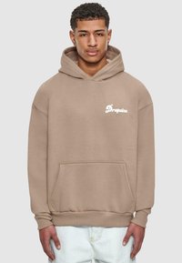Beige hettegenser med lommer foran og "Dropsize"-logo i hvitt. Har en løs passform og ribbede mansjetter. Brukt over lysejeans.