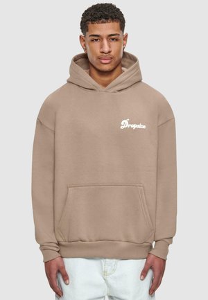 Beige hettegenser med lommer foran og "Dropsize"-logo i hvitt. Har en løs passform og ribbede mansjetter. Brukt over lysejeans.