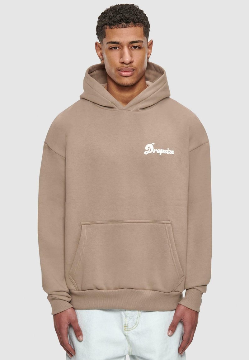 Beige hettegenser med lommer foran og "Dropsize"-logo i hvitt. Har en løs passform og ribbede mansjetter. Brukt over lysejeans.