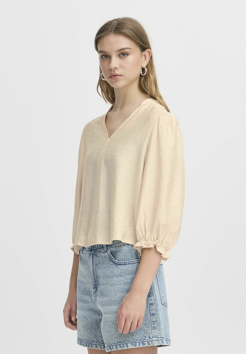 Ljus beige blus i ett mjukt tyg, med V-ringning, långa puffärmar med volangärmar och en lös passform, i kombination med denimshorts.