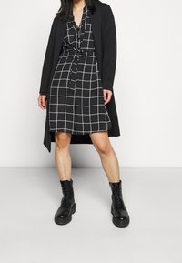 Černý dlouhý kabát přes černo-bílou kostkovanou šaty s páskem v pase, doplněný černými šněrovacími kotníkovými botami. Plochá podrážka, texturovaný povrch.