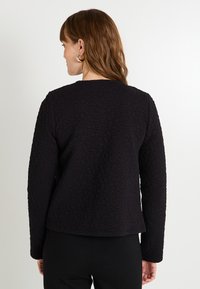 Chaqueta negra texturizada con patrones florales, cuello redondo y mangas largas. La tela parece suave y ligeramente estructurada.