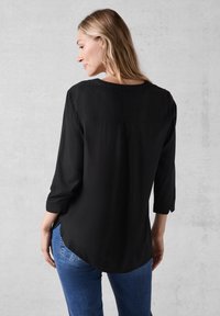 Sort, let og luftig bluse med rund halsudskæring, trekvartærmer og en plissering bagpå. Båret med blå denim jeans.