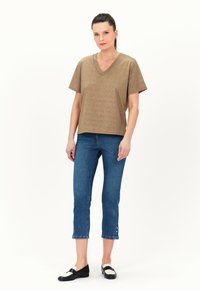 Bronzefarbenes T-Shirt mit kurzen Ärmeln und V-Ausschnitt in lockerer Passform, kombiniert mit blauen knöchellangen Jeans mit Knopfdetails am Saum und schwarzen Ballerinas.
