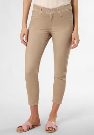 Femme portant un pantalon beige slim-fit coupé et des sandales plates ouvertes roses, se tenant devant un fond blanc uni.