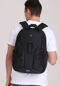 Sac à dos noir avec bretelles rembourrées, plusieurs compartiments et accents orange. Texture de tissu lisse, design épuré, adapté à un usage quotidien.